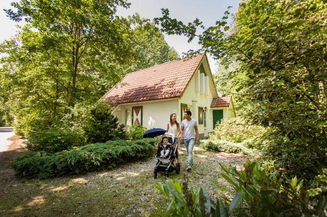 Ein Häuschen im Ferienpark, an dem eine junge Familie mit einem Kinderwagen vorbeigeht.